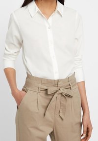 White button-up shirt with a classic collar and long sleeves paired with high-waisted beige trousers featuring a tied waist and front pockets.