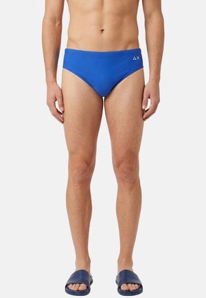 Uomo che indossa slip da bagno blu e sandali navy, in piedi contro uno sfondo chiaro e uniforme.