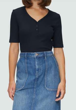 Kvinde iført en navy kortærmet henley-top, der er stoppet ned i et højtaljet blåt denimsnørk med store forlommer.