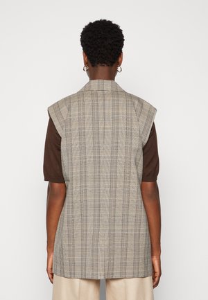 Osoba o krótkich kręconych włosach, nosząca złote kołowe kolczyki, bezrękawnik w kratę na brązowej bluzce z krótkim rękawem i beżowe spodnie, zwrócona tyłem.