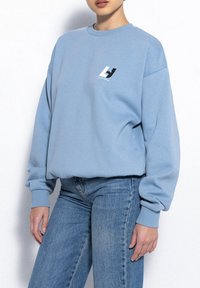 Jasnoniebieski sweter z ribbowanymi mankietami i okrągłym dekoltem. Na klatce piersiowej znajduje się małe czarno-białe logo. W zestawie z niebieskimi dżinsami.