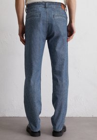 Niebieskie dżinsowe spodnie o prostym kroju, z kieszonką w pasie, tylnymi kieszeniami oraz subtelną teksturowaną powierzchnią. Noszone z czarnymi butami.