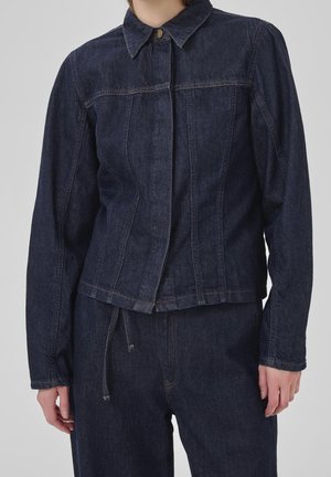 Vrouw met een donkerblauwe spijkerjas met kraag en bijpassende spijkerbroek tegen een effen achtergrond.
