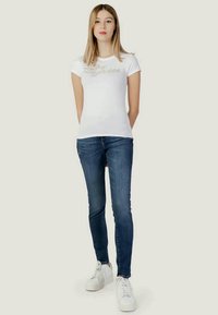 Bijela majica s glitternim "Guess" logom, uparena s tamnoplavim slim-fit trapericama i bijelim tenisicama. Jednostavan dizajn i prilagođeni kroj.