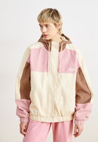 Farvet blokket corduroy jakke med creme-, pink- og brune sektioner. Inkluderer justerbar hætte og elastiske manchetter. Roxy-logoet er til stede.