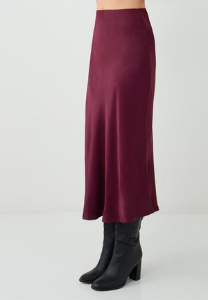 Burgunderfarbene Satin-Midirock mit glatter Textur, hoch geschnittenem Bund und fließender Silhouette, kombiniert mit schwarzen Stiefeln mit Absatz.