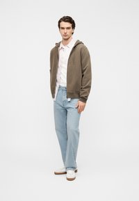 Γκρι φούτερ με φερμουάρ πάνω από ροζ ανοιχτό πουκάμισο, συνδυασμένο με ανοιχτό μπλε τζιν και λευκά αθλητικά παπούτσια. Το σύνολο χαρακτηρίζεται από ουδέτερους τόνους και casual στυλ.