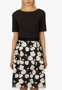 Schwarzes Kleid mit kurzen Ärmeln, figurbetontem Oberteil und vollem Rock, der mit weißer Blumen-Spitze auf schwarzem Hintergrund überzogen ist.