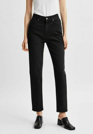 Femme portant un jean droit taille haute noir associé à des mules noires à talons et un top sans manches blanc sur un fond uni.