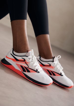 Persona con zapatillas deportivas Reebok blancas, negras y naranja neón, y leggings negros, de pie sobre un suelo de color claro.