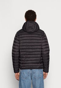 Mężczyzna w czarnej pikowanej kurtce z kapturem i niebieskich dżinsach, stojący tyłem do widza na jednolitym tle.