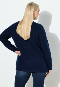 Blondynka z prostymi włosami, nosząca granatowy sweter i ciemne dżinsy, stoi z jedną ręką na biodrze, przodem do białej ściany.