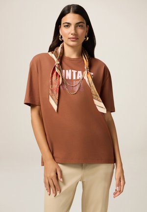 Femme aux longs cheveux noirs portant des boucles d'oreilles en or, un t-shirt marron "Vintage Soul", un foulard à motifs noué comme un collier, et un pantalon beige.