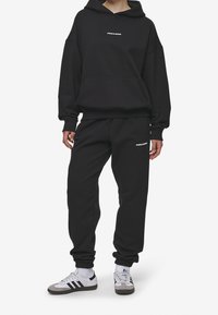 Sudadera de algodón negra con un bolsillo frontal y logotipo, combinada con pantalones de chándal negros a juego que tienen puños elásticos. Llevada con zapatillas blancas y grises.