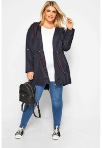 Laivaston vedenkestävä takki säädettävällä vyötäröllä, vetoketjulliset taskut ja punaisia yksityiskohtia. Yhdistetty valkoiseen paitaan, sinisiin farkkuihin ja mustaan reppuun.