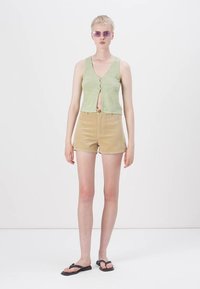 Green sleeveless top with a front button detail, paired with beige corduroy shorts. Model wears thin, purple sunglasses and black flip-flops.