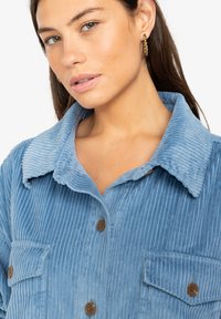Camicia di velluto a coste azzurro chiaro con colletto pieghevole, chiusura con bottoni e due tasche sul petto. Tessuto a coste testurizzato con bottoni marroni.