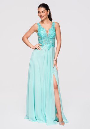 Femme portant une robe aqua sans manches avec corsage en dentelle, décolleté en V profond, fente haute sur la cuisse et talons argentés, souriante avec la main sur la hanche.