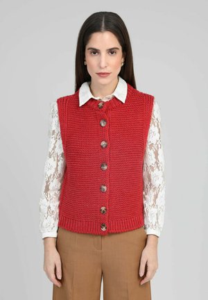 Gilet rouge tricoté à texture côtelée, avec un col rond et une fermeture avant à boutons en bois, porté sur une chemise blanche à manches longues en dentelle.