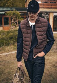 Brązowa ocieplane kamizelka noszona na granatowej koszuli z długim rękawem, w połączeniu z ciemnymi dżinsami i beżową torebką na ramię. Swobodna sceneria na świeżym powietrzu.