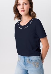 Marineblaues Kurzarmoberteil mit Rundhalsausschnitt. Mit silbernen Akzenten am Ausschnitt. Kombiniert mit hellblauen, verzierten Jeans.