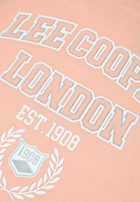 Sweat-shirt couleur pêche avec texte en blanc et gris clair indiquant "LEE COOPER LONDON EST. 1908", avec un détail de blason et de lauriers.