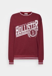 Sudadera burdeos con texto "Hollister" impreso en blanco y diseño de laurel. Cuenta con puños y cuello acanalados con franjas blancas. Material suave.