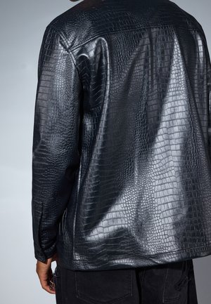 Mann trägt eine glänzende schwarze Jacke aus Kunst-Krokodilleder, von hinten vor einem einfarbigen grauen Hintergrund zu sehen.