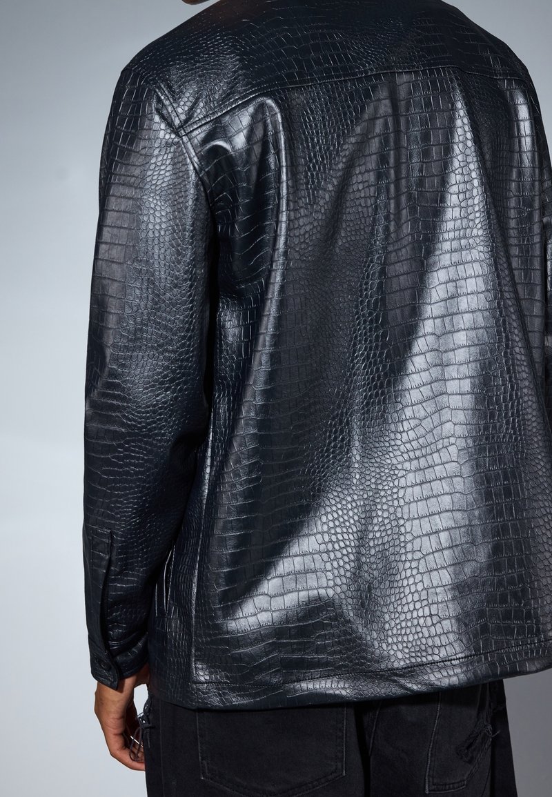 Homme portant une veste noire brillante en cuir synthétique effet crocodile, vu de dos sur un fond gris uni.