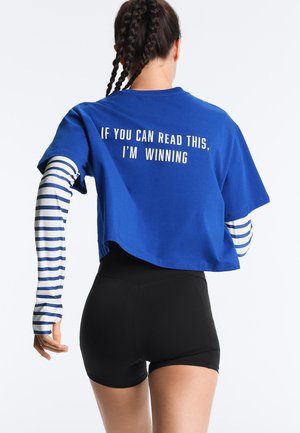 Donna con capelli intrecciati che indossa una T-shirt corta blu con la scritta "IF YOU CAN READ THIS, I'M WINNING", pantaloncini neri e maniche lunghe a righe.