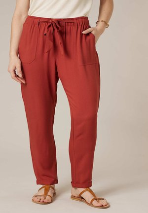 Vrouw die roestkleurige broek met trekkoord en zakken draagt, gecombineerd met tankleurige sandalen en een crèmekleurige top, staand met één hand in de zak.