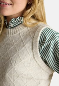Frau trägt einen cremefarbenen, gestrickten, ärmellosen Pullover über einer grün-weißen, gestreiften Bluse mit Rüsche am Kragen und Perlenohrringen.