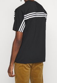 Camiseta negra con tres franjas blancas horizontales en la parte posterior y las mangas. Hecha de algodón, tiene un escote redondo y mangas cortas.