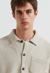 Giacca camicia in tessuto beige chiaro con colletto, bottoni neri, taschino frontale e motivo in tessuto intrecciato.