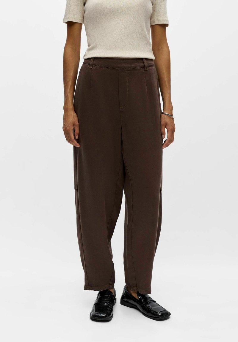 Femme portant un pantalon marron ample, un haut beige à manches courtes, des mocassins noirs, debout devant un fond blanc uni.