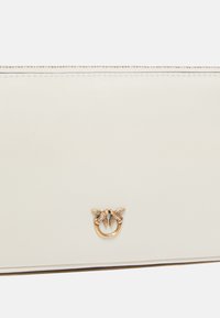 Portafoglio in pelle crema con chiusura a zip, caratterizzato da un emblema dorato e dettagli accurati. Texture liscia, stile minimalista.