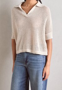 Jasnobeżowy krótki sweter z krótkim rękawem i kołnierzykiem, o luźnym kroju i otwartym wzorze dzianiny, zestawiony z jasnoniebieskimi jeansami o prostych nogawkach.