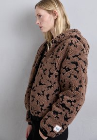 Brown, fuzzy zip-up jacket with black abstract pattern, elastic cuffs, and a hood. Label with "EA7" visible at the hem.