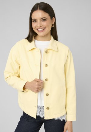 Femme souriante portant une veste boutonnée jaune pâle sur un haut blanc avec un ourlet en dentelle et un jean foncé, debout devant un fond gris.