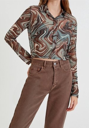 Femme portant un crop top boutonné semi-transparent à manches longues avec des motifs tourbillonnants marron, bleu et crème, associé à un pantalon taille haute marron.