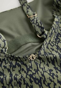 Gros plan sur un tissu à motifs verts et noirs avec des bretelles réglables et un petit détail de fermoir circulaire doré.