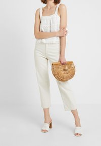 Top blanco sin mangas con detalle de ojal, pantalones vaqueros de pierna ancha color crema, sandalias deslizantes blancas de tejido, y un bolso de bambú en forma de media luna.