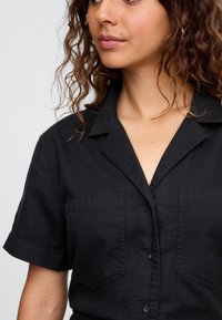 Camisa negra de manga corta en un tejido ligero, con cuello, dos bolsillos en el pecho y cierre delantero con botones. Textura lisa, diseño casual.