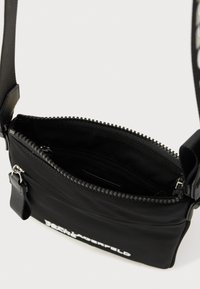 Czarna torba na pasku z poliamidu z zamkiem błyskawicznym, kieszeniami wewnętrznymi i regulowanym paskiem. Na froncie znajduje się nadrukowane logo. Gładka faktura.