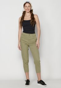 Pantalones cortos de color verde oliva con cierre de botón en la parte delantera y presillas para el cinturón, combinados con una camiseta sin mangas negra y zapatillas negras. La modelo está de pie contra un fondo blanco.