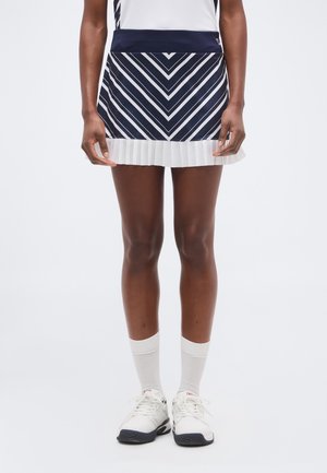 Vrouw gekleed in een tennisrok met een marineblauw-wit zigzagpatroon en een witte geplooide zoom, witte sokken en witte tennisschoenen, rechtstaand.