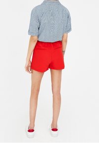 Shorts rouges à la texture lisse, à la coupe ajustée et dotés de deux poches arrière, assortis à une chemise à col à rayures. Chaussures blanches à enfiler.