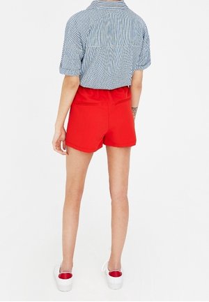 Shorts rouges à la texture lisse, à la coupe ajustée et dotés de deux poches arrière, assortis à une chemise à col à rayures. Chaussures blanches à enfiler.