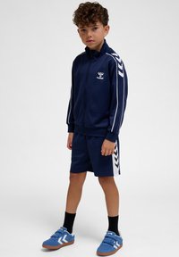 Jongen staand, gekleed in een marineblauw sportjack en bijpassende korte broek met witte strepen, zwarte sokken en blauwe sneakers tegen een effen achtergrond.