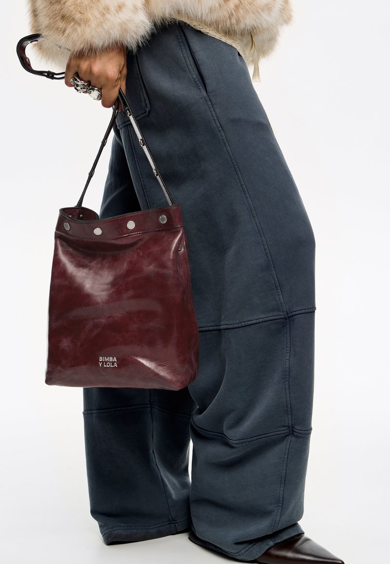 Czerwona skórzana torba typu bucket z srebrnym okuciem i logo, trzymana przez osobę w luźnych, ciemnoniebieskich, teksturowanych spodniach.